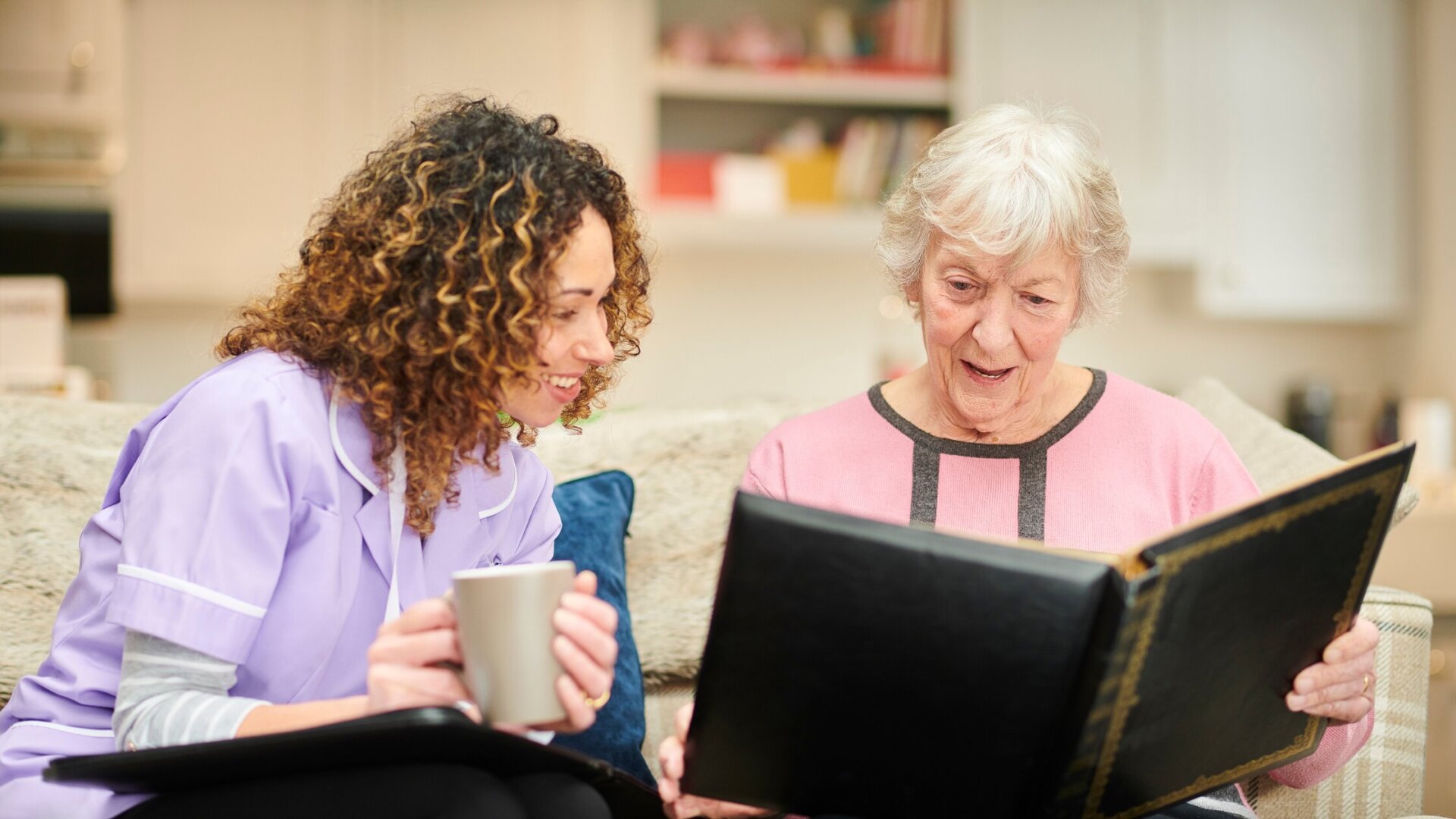 Senior woman sitting on the couch with her caregiver while looking through old photos at Traditions at Hunter Station