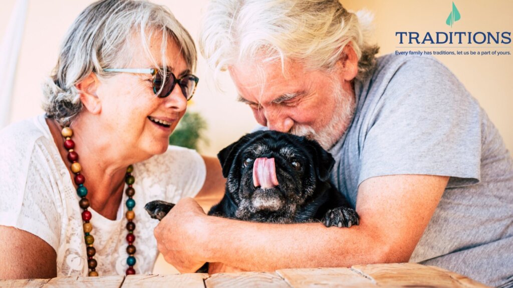 An elderly couple is hugging a black pug and smiling with the Traditions logo in the top right corner.